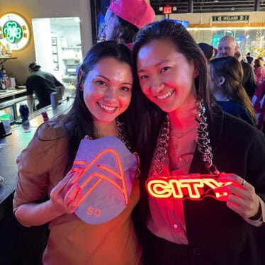 Two women smiling at a bar, each holding large, glowing signs around their necks—one with a colorful geometric design and the other with the word "CITY" in bright neon letters. People and bar decor are visible in the background.